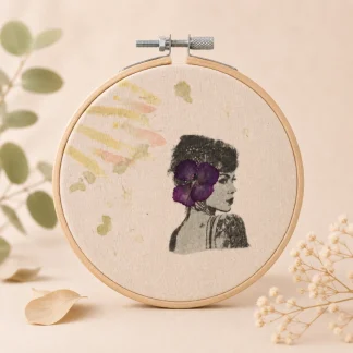 Vista frontal de un bastidor circular con retrato de perfil en negro, flor morada y estampaciones botánicas sobre tela